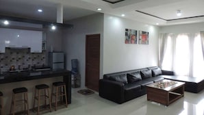 A modern living room with a black leather sofa, a coffee table, and a kitchen area with a bar counter and stools. - Villa Fahim 1 Puncak 4 Bedroom (Cipanas)