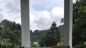 A wooden outdoor seating area with a table and chairs, a view of a lush garden and trees, and a cloudy sky. - Villa Fahim 1 Puncak 4 Bedroom (Cipanas)