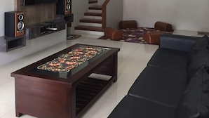 A modern living room with a flat-screen TV, wooden entertainment unit, a coffee table with decorative items, a black sofa, and a staircase leading to another level. - Villa Fahim 1 Puncak 4 Bedroom (Cipanas)