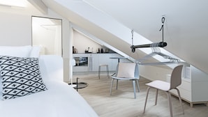 A modern loft bedroom with a sloped ceiling, a bed with patterned pillows, a desk with a chair, and a kitchenette in the background. - Dom Studios (St. Gallen)