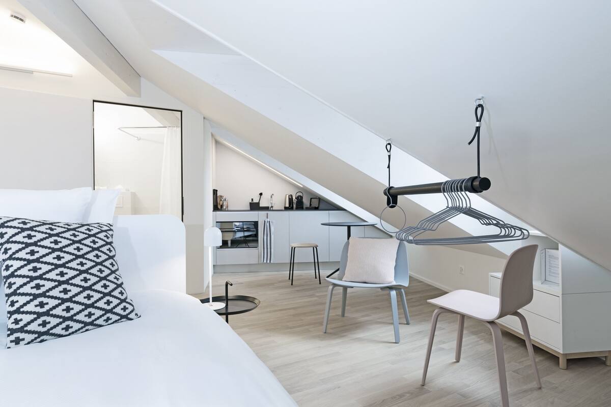 A modern loft bedroom with a sloped ceiling, a bed with patterned pillows, a desk with a chair, and a kitchenette in the background.