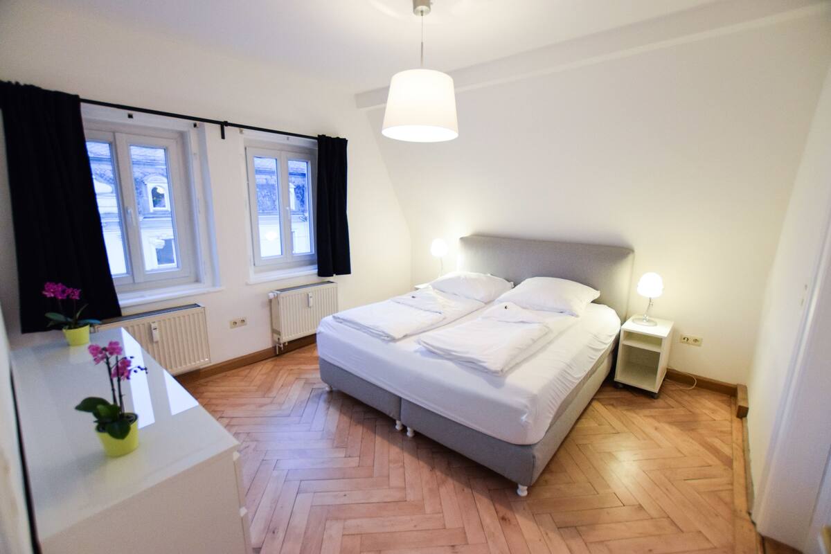 Ein Schlafzimmer mit Bett, Nachttischen, einem Schreibtisch mit Pflanze und einem fenster mit Vorhängen.