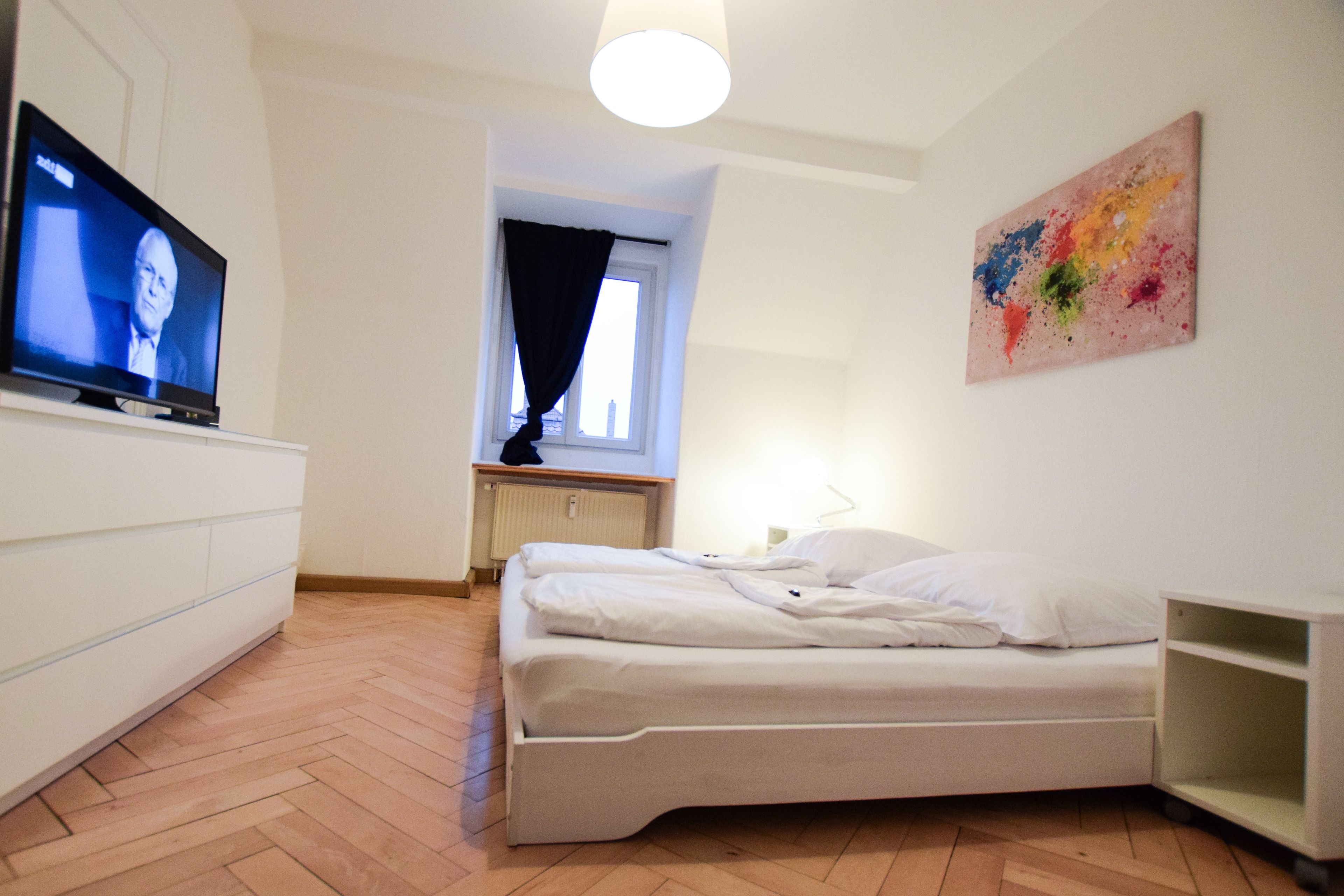 Ein Schlafzimmer mit einem Bett, einem Fernseher auf einer Kommode, einem Fenster mit Vorhängen und einem farbenfrohen Bild an der Wand.