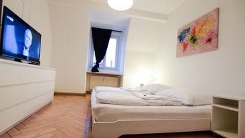 Ein Schlafzimmer mit einem Bett, einem Fernseher auf einer Kommode, einem Fenster mit Vorhängen und einem farbenfrohen Bild an der Wand.