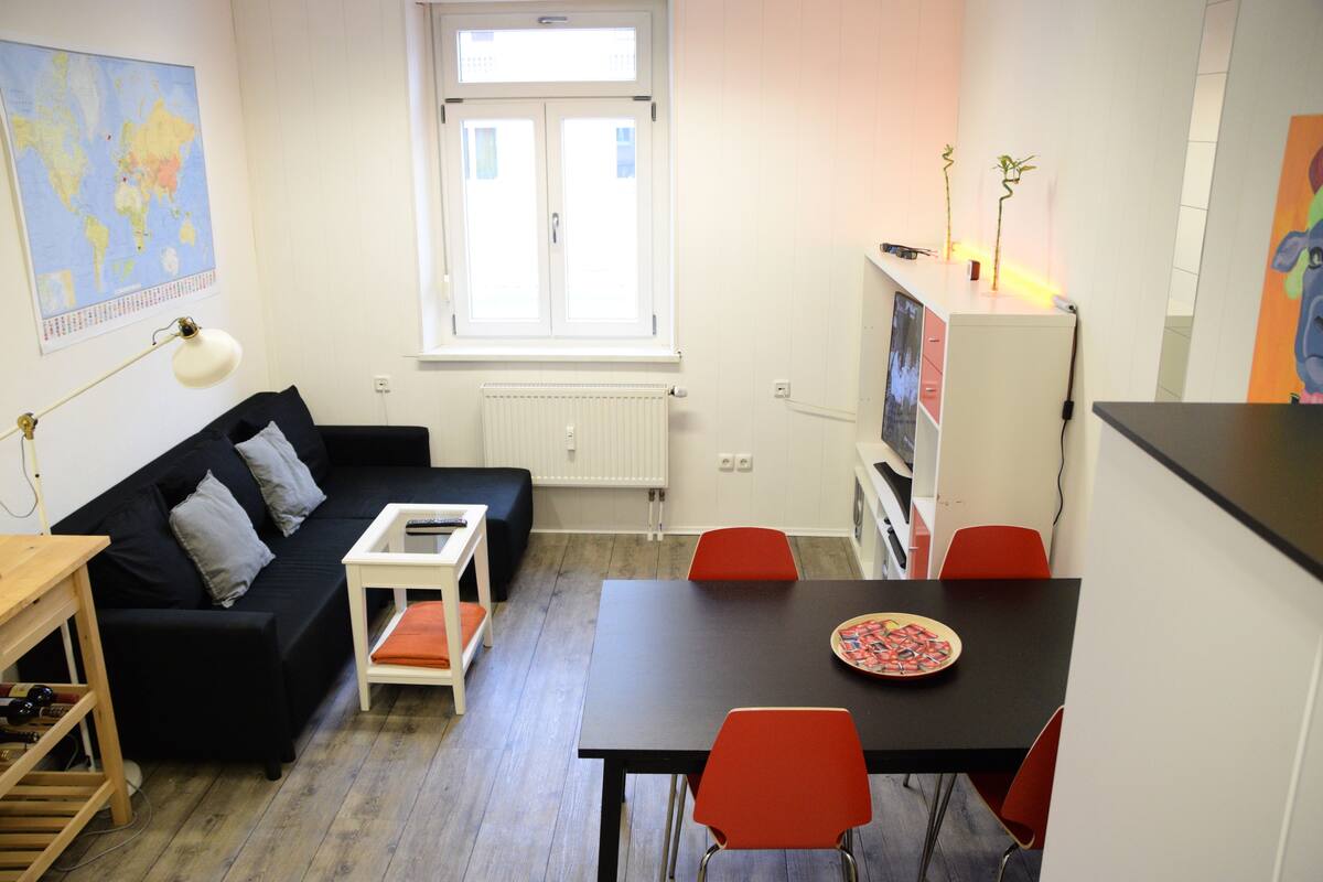 Ein kompakter Wohnraum mit schwarzem Sofa, kleinem weißen Tisch, einem schwarzen Esstisch mit roten Stühlen, einem weißen Schrank und einer Weltkarte an der Wand.