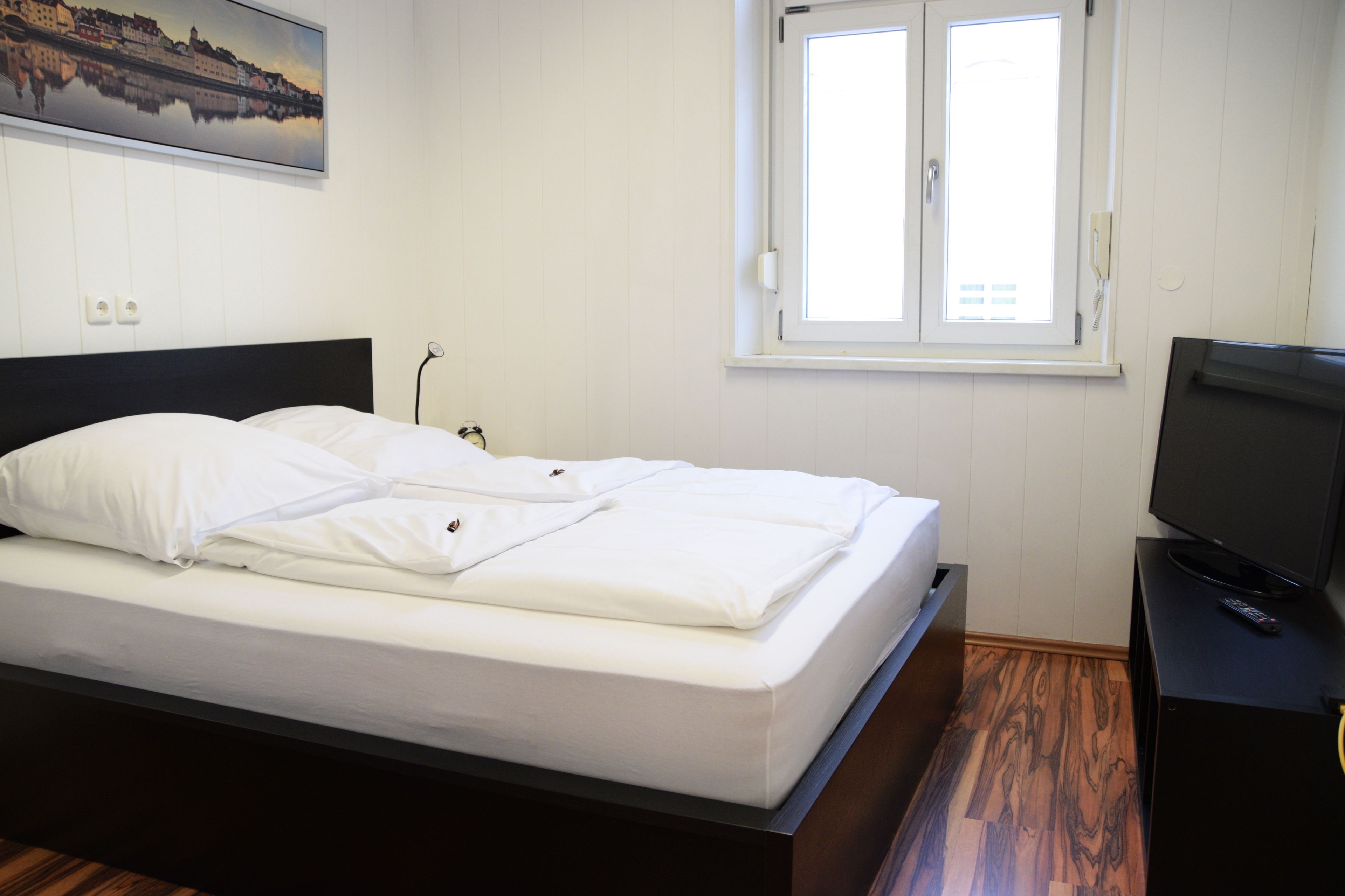 Ein ordentlich bezogenes Bett mit weißen Laken, ein schwarzes Kopfende, ein Fenster mit weißen Rahmen und ein Fernseher auf einem Holzständer.