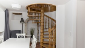 Uma escada em espiral em um espaço com uma mesa e cadeiras brancas, um abajur e uma planta.