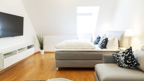 Uma sala de estar moderna com televisão de tela plana, um sofá cinza, uma mesa pequena com abajur e uma janela que permite entrada de luz natural.