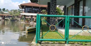 A lakeside view with a boat docked near a green metal fence, a stone pillar, and a building with a tiled roof. - Villa Pinggir Danau Puncak for 30 pax (Cipanas)