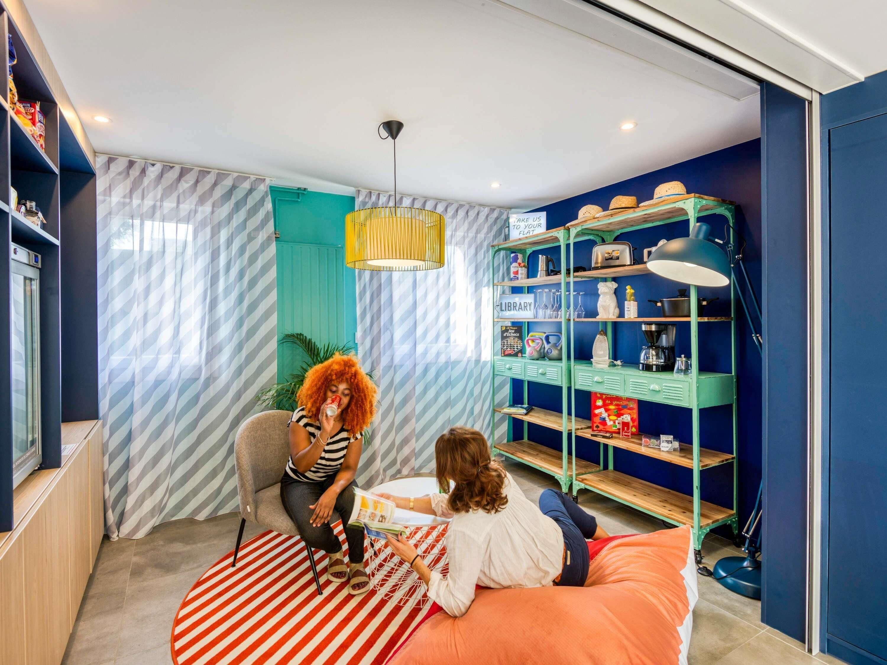 Een kamer met een muur in teal, een gestreept gordijn, een groen metalen rek met het opschrift 'BIBLIOTHEEK' en een rood-wit gestreept vloerkleed.