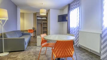 A modern hotel room with a round dining table, orange chairs, a grey sofa, a TV mounted on the wall, and a closet with a mirror.