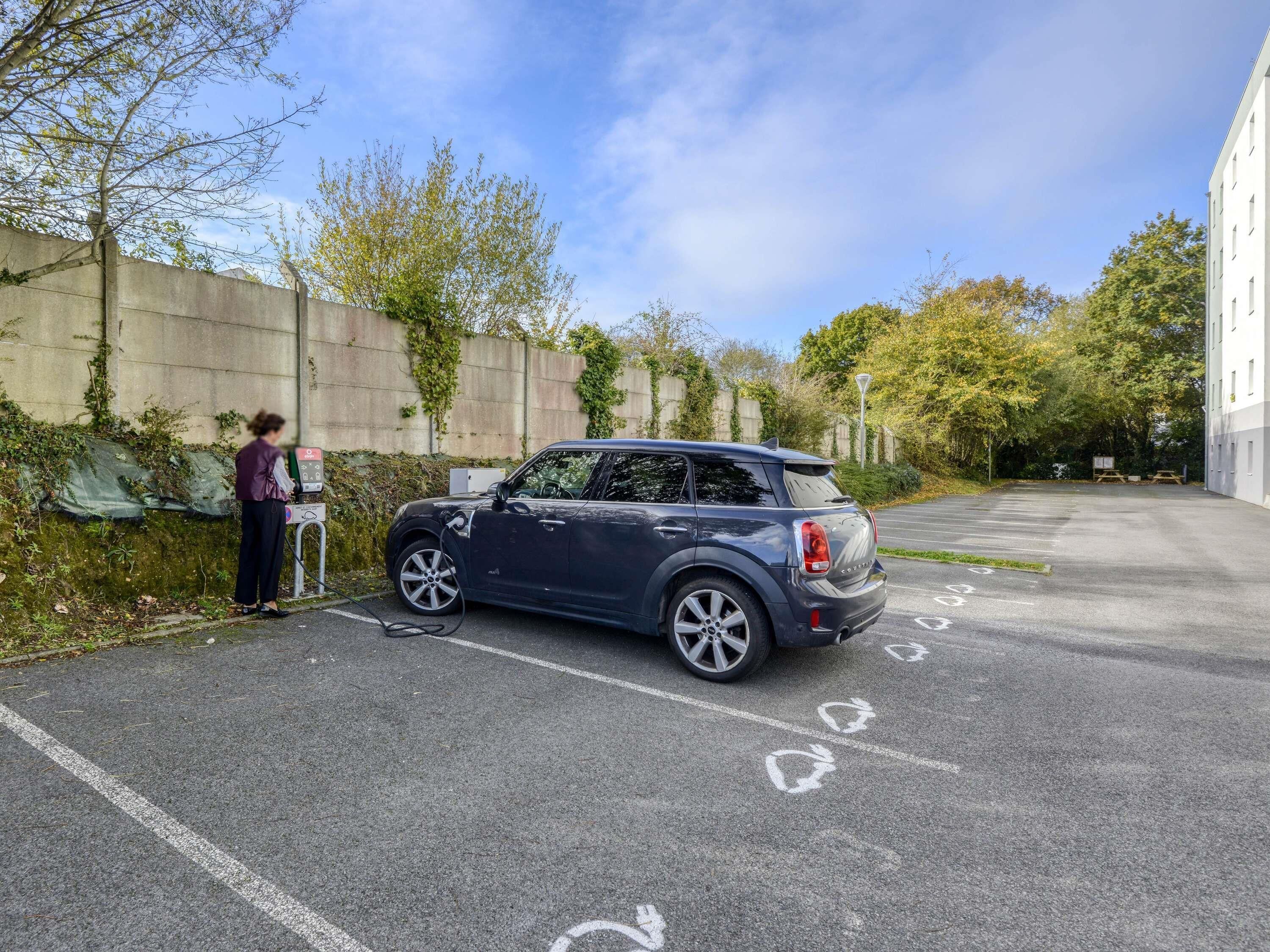 A parking area with a Mini Cooper parked and charging, a person nearby, and a building in the background.