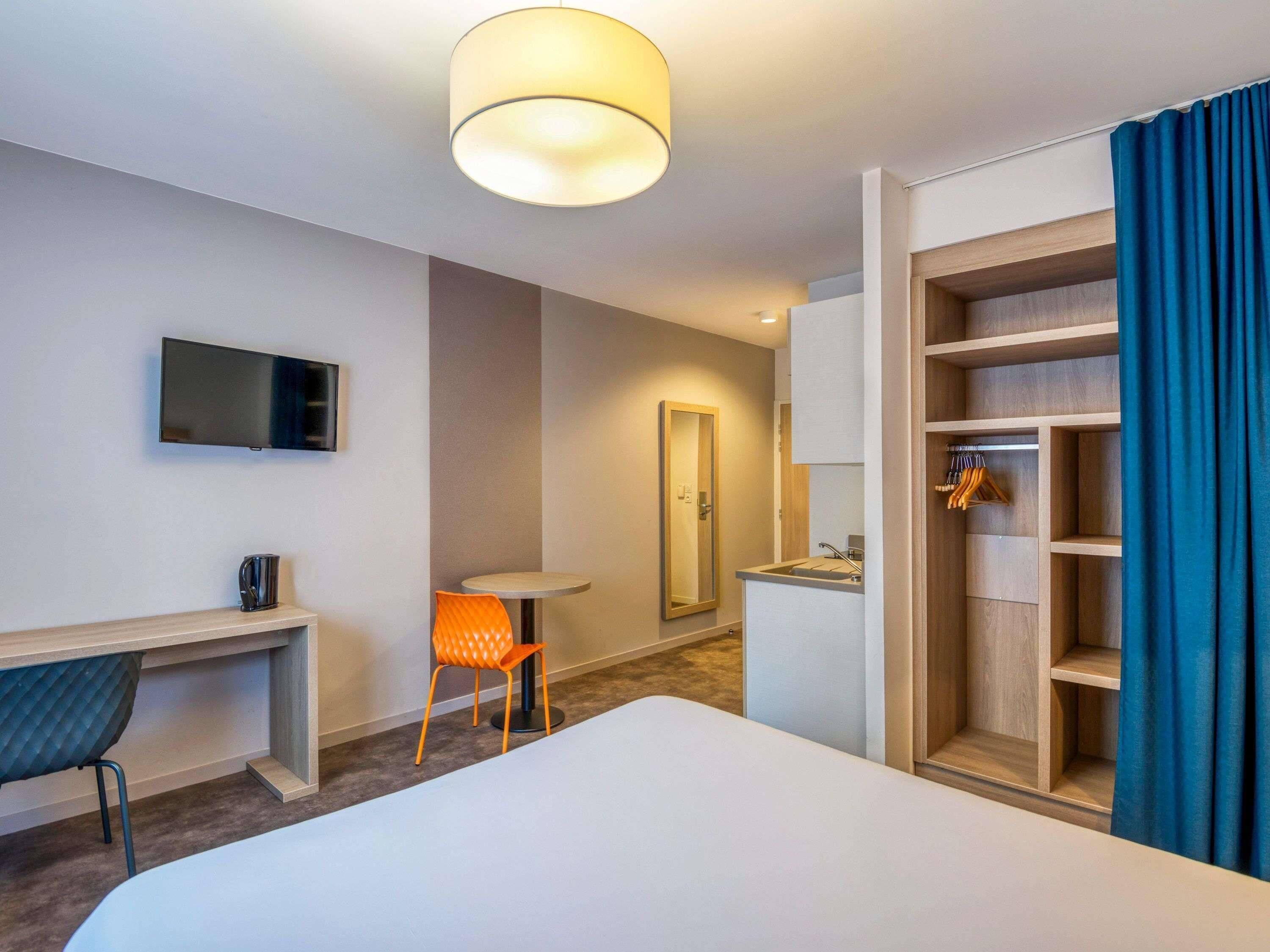 A hotel room with a bed, a desk, a chair, a TV, a mirror, a wardrobe, and a bathroom with a sink and mirror.