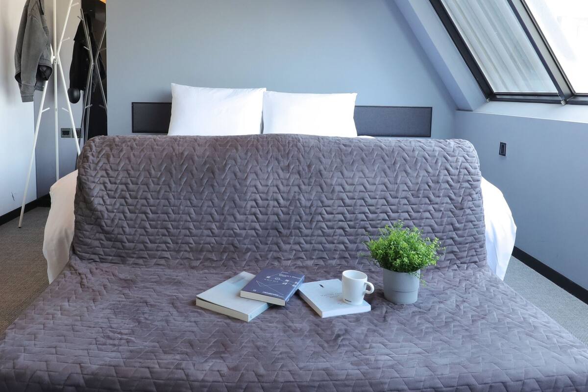 A neatly made bed with a grey quilt, a small potted plant, and a couple of books and a mug on the bed.