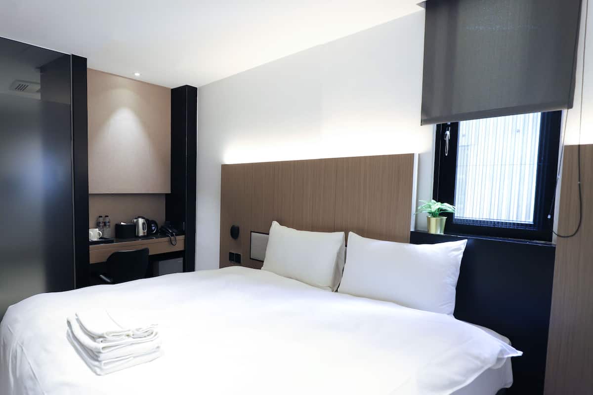 A modern hotel room with a neatly made bed, a small desk with a coffee maker, and a window with blinds.