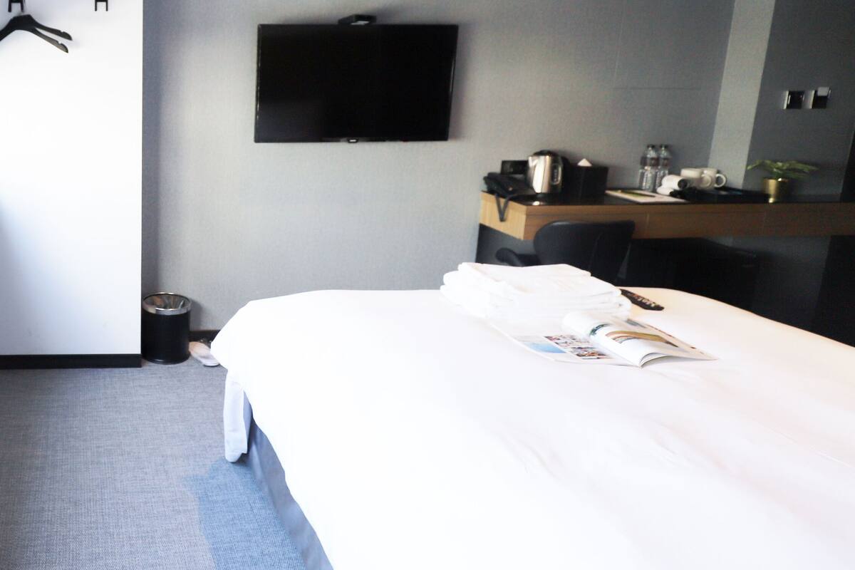 A hotel room with a bed, a TV mounted on the wall, a small table with a kettle, and a window with blinds.