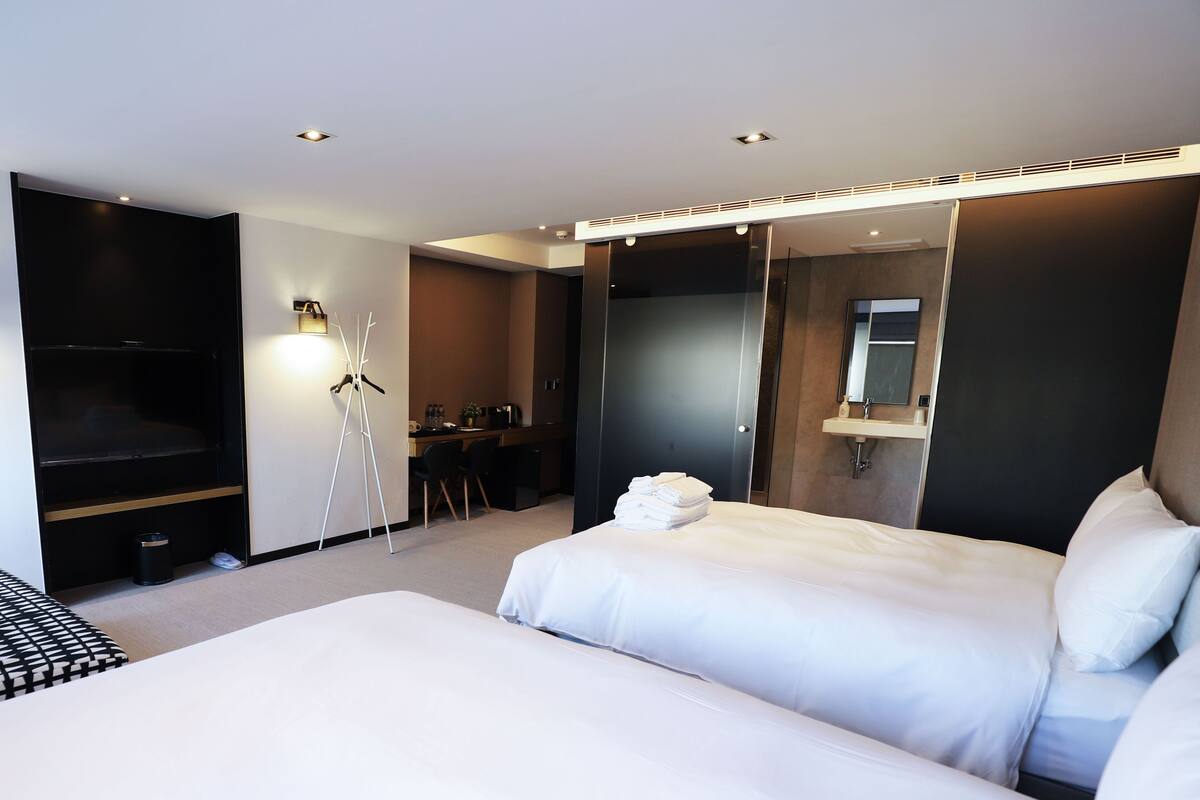 A modern hotel room with a large bed, a desk with a chair, a TV, and a bathroom with a mirror.