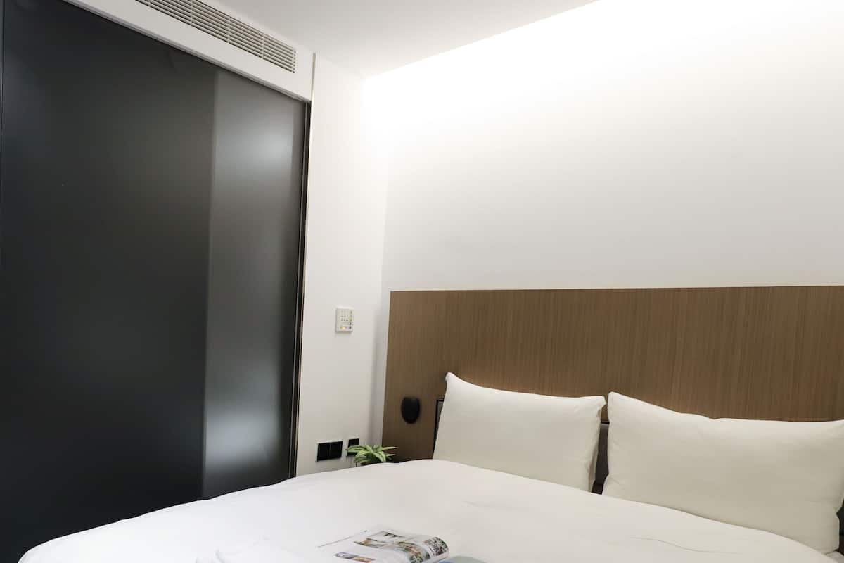 A hotel room with a bed, a control panel, a plant, and a wall-mounted air vent.