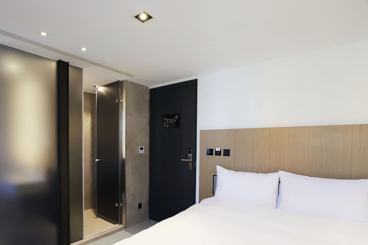 A modern hotel room with a large bed, a wooden headboard, a dark door, and a glass shower enclosure.