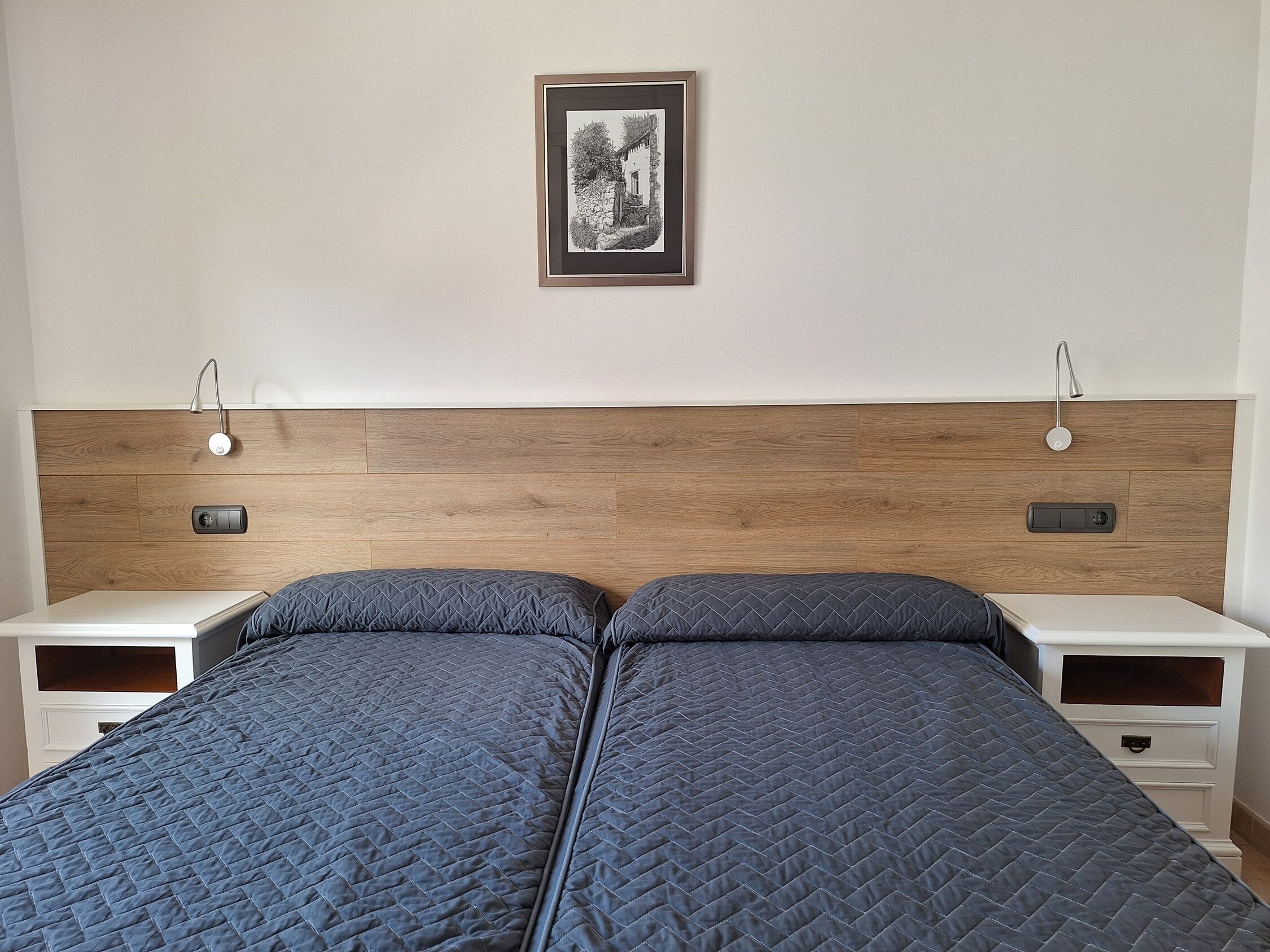 Una cama doble con cabecera acolchada, dos mesitas de noche con lámparas, y un cuadro en la pared.