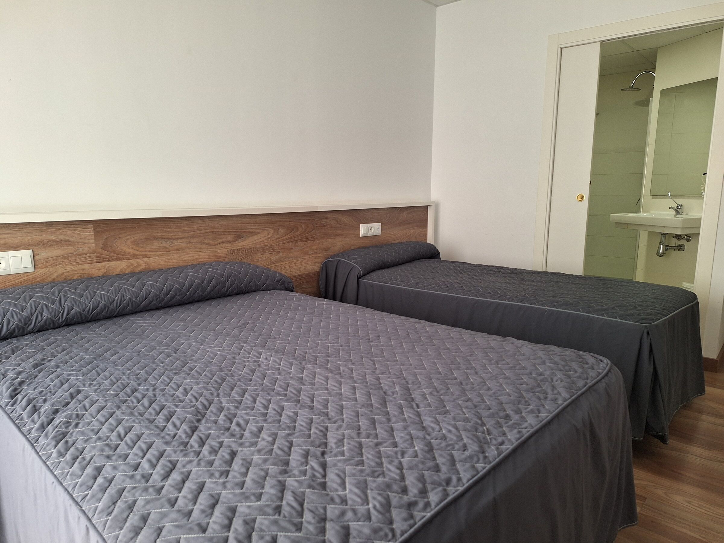 A room with two beds, a wooden headboard, a white wall, and a bathroom visible through an open door.