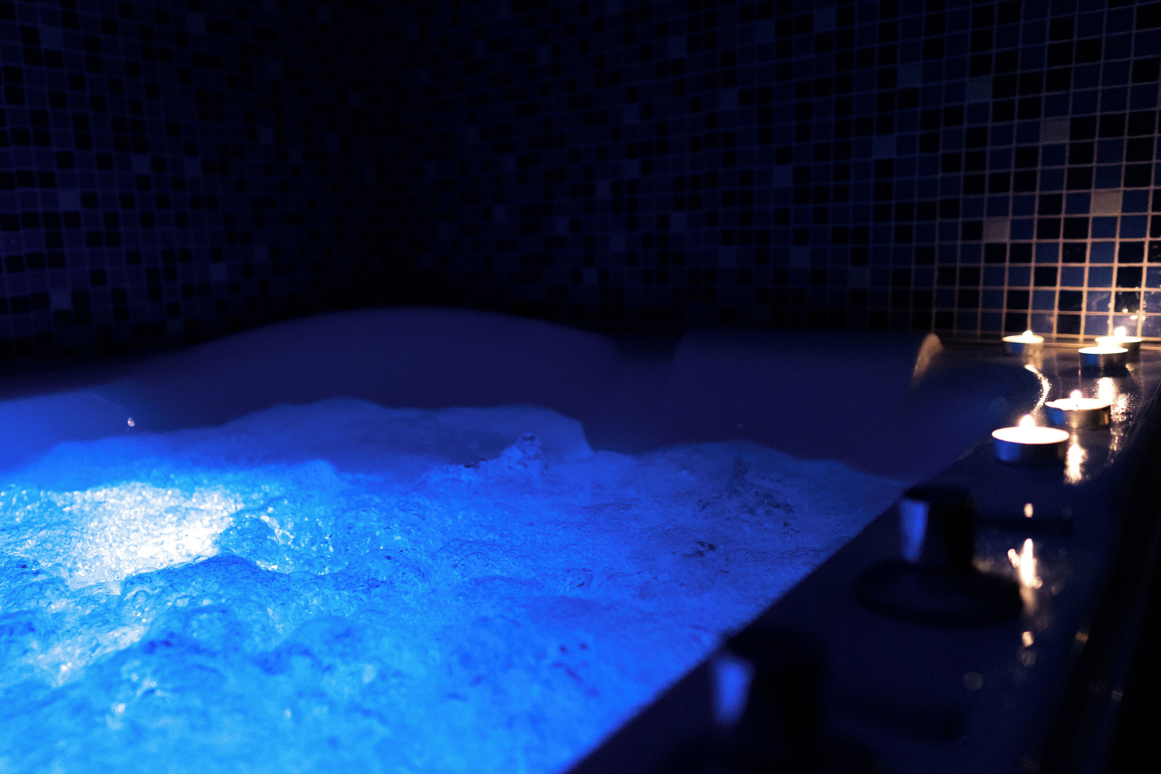 A bathtub with blue lighting, a tiled wall, and candles.