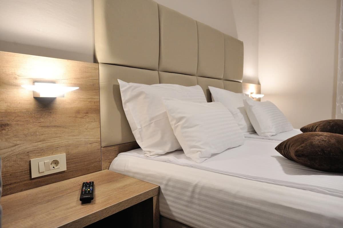 A hotel room with a bed, white pillows, a remote control on a wooden nightstand, and wall-mounted lights.