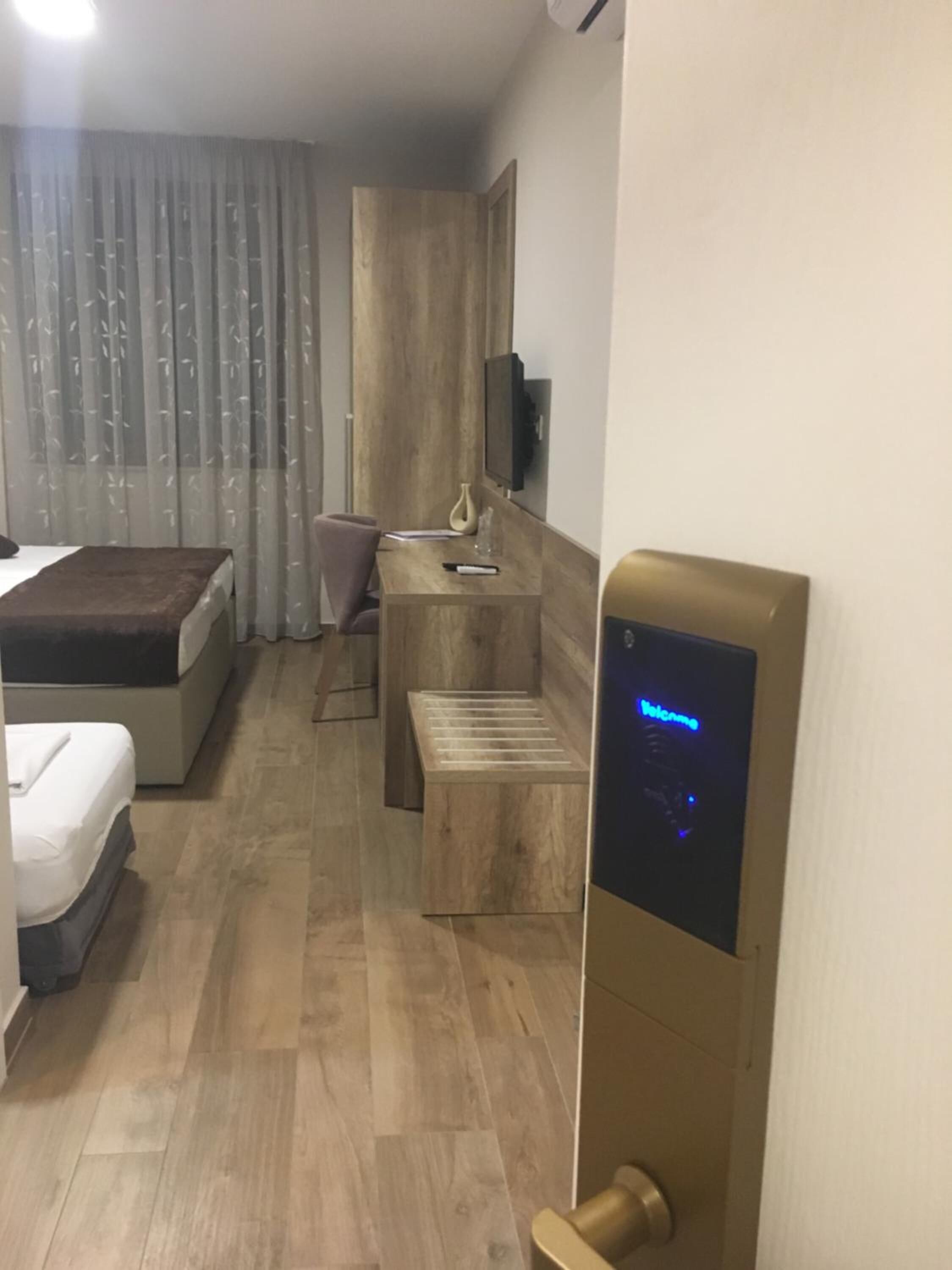 A hotel room with a bed, a desk, a chair, a small table, a TV mounted on the wall, and a digital lock on the door.