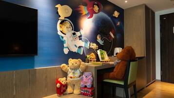 A room with a wall mural of astronauts and space-themed decorations, a flat-screen TV, and a desk with a teddy bear and a small table with a toy car.