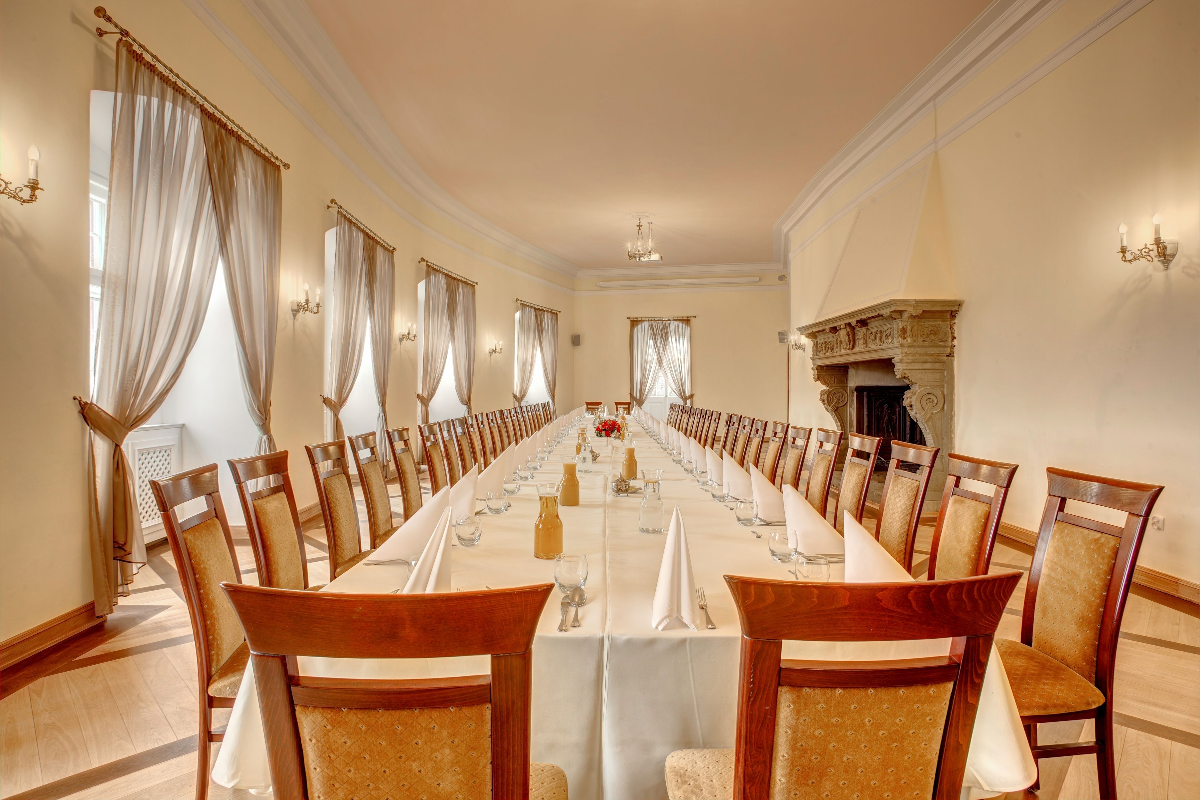 A long dining table set for a formal meal, flanked by wooden chairs with cushioned seats, in a room with cream walls and a fireplace.