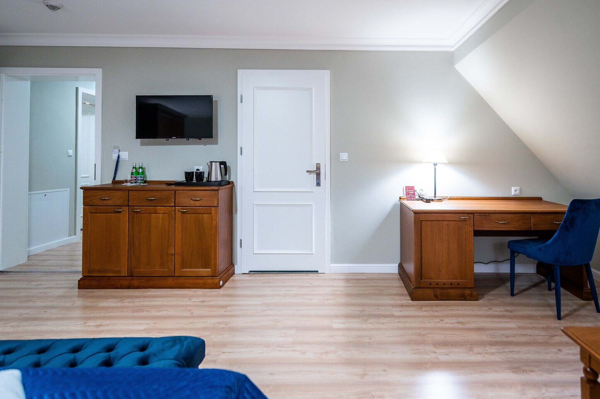A room with a wooden desk, a blue chair, a television mounted on the wall, and a wardrobe.