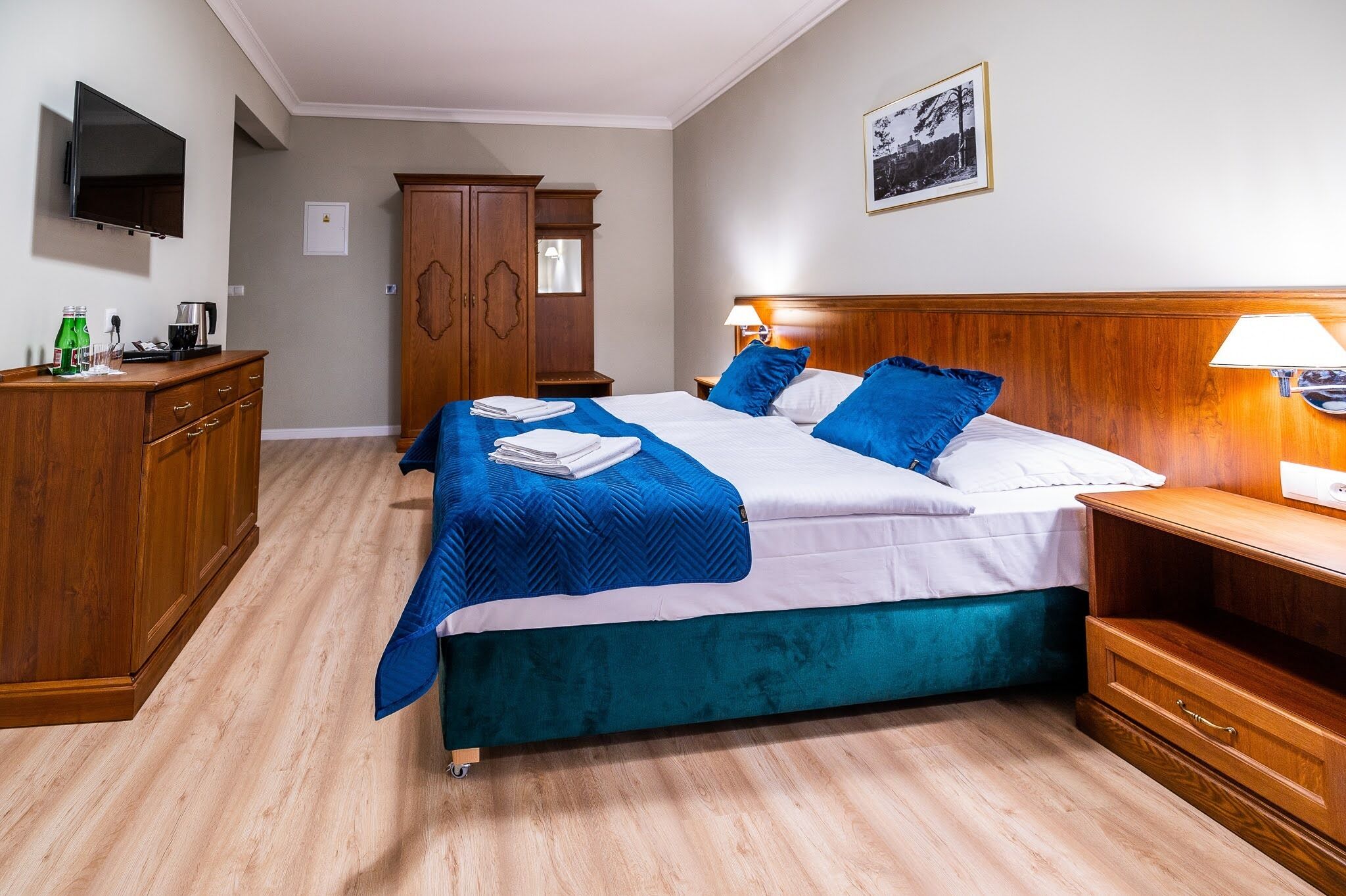 A hotel room with a bed, a wooden wardrobe, a TV mounted on the wall, and a framed picture on the wall.