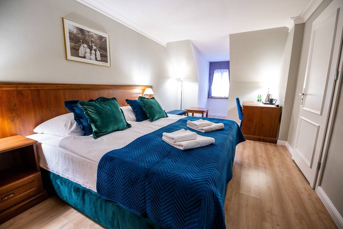 Ein ordentlich bezogenes Bett mit blauer und weißer Bettdecke, ein Nachttisch aus Holz, ein Beistelltischchen mit einer Lampe und ein gerahmtes Foto an der Wand.