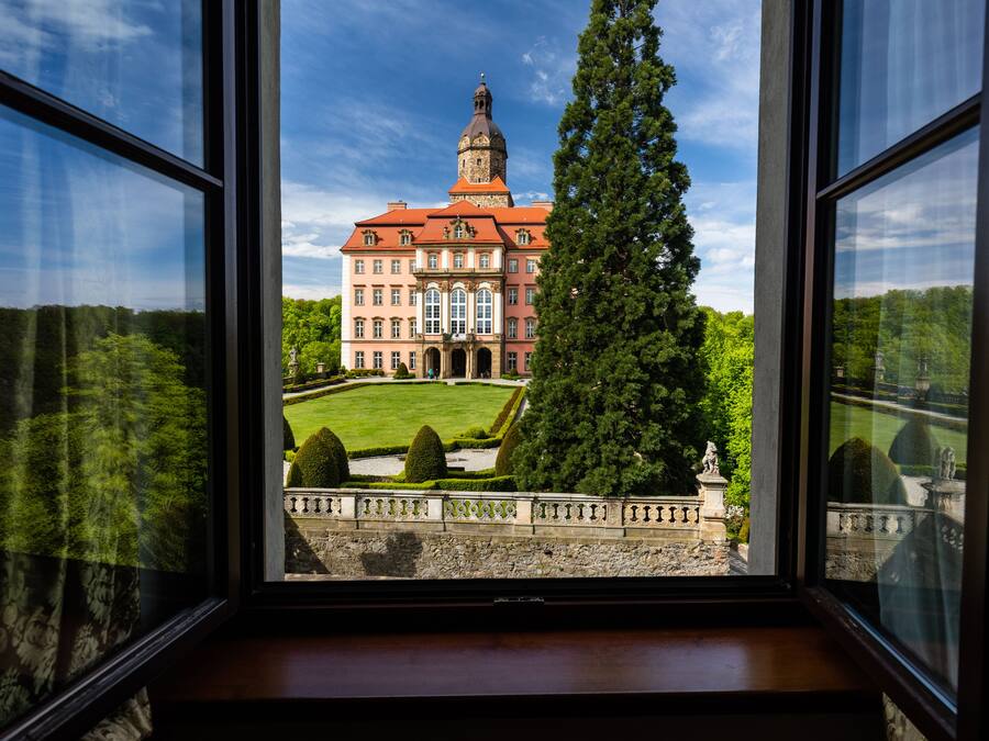 A large, historic building with a prominent tower, viewed through a window with a stone balustrade.