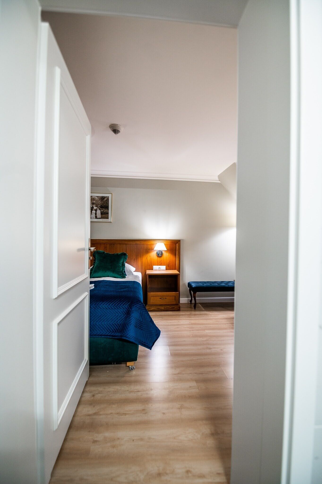 Een slaapkamer met een bed, nachtkastje, lamp en een bankje.