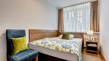 Habitación de hotel con cama, una silla, una mesita de noche y un ventanal con cortinas.