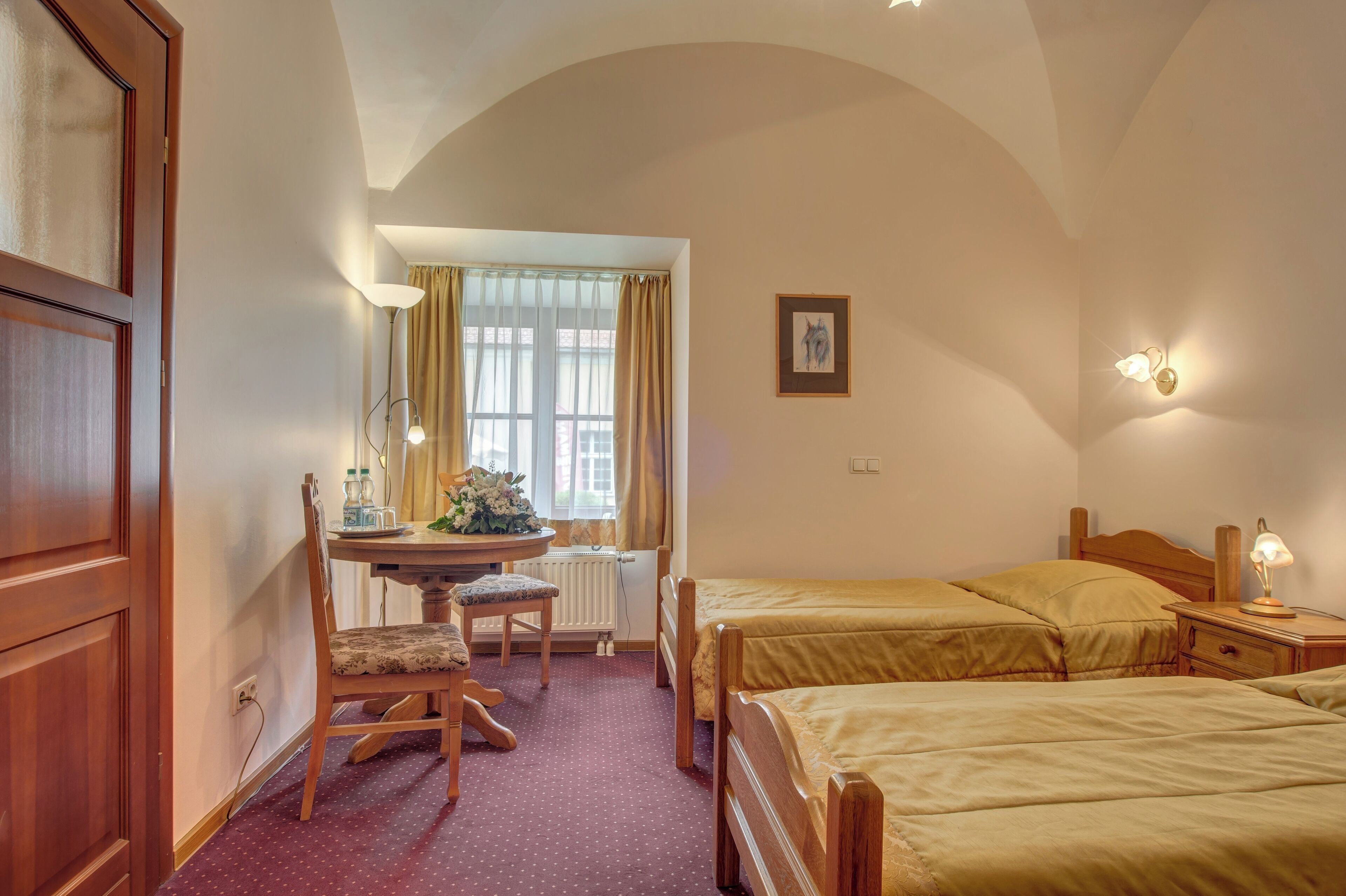 A hotel room with two single beds, a wooden desk with a chair, a radiator, and a window with curtains.