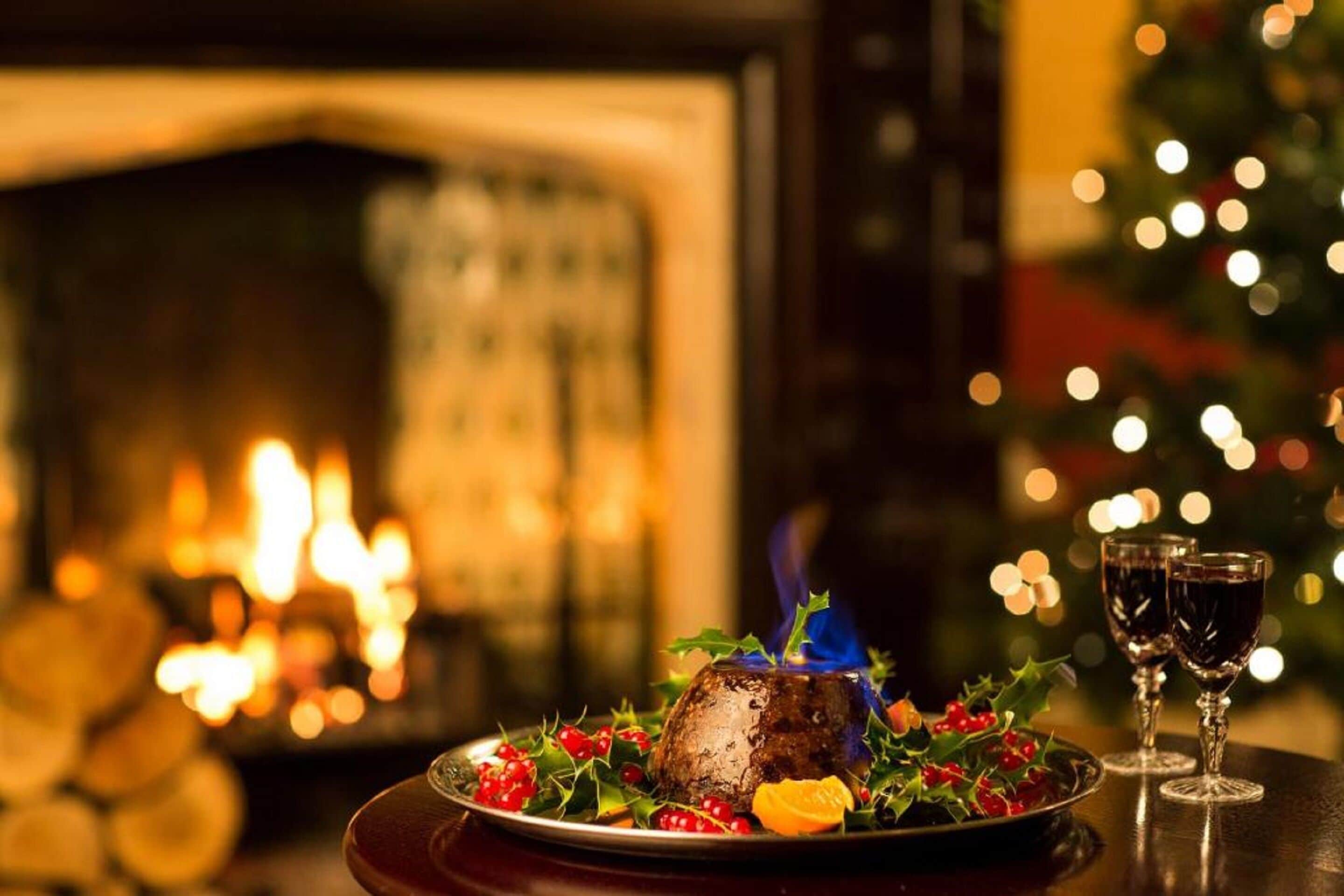 A Christmas pudding on a plate with holly and berries, two wine glasses, and a lit fireplace in the background.