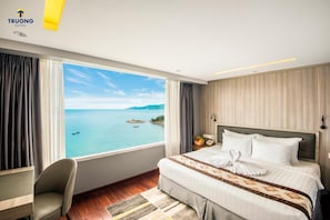 A hotel room with a large window offering a view of the sea and a small island. The room has a bed with white linens, a grey armchair, and a wooden floor.