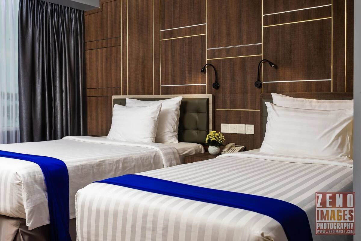 Une chambre d’hôtel avec deux lits, une tête de lit en bois, une table de chevet avec un téléphone et une fenêtre avec des rideaux.
