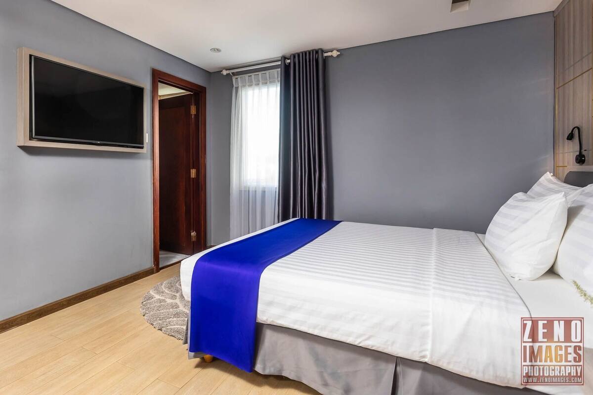 Une chambre d’hôtel avec un lit, une télévision, une fenêtre avec des rideaux et une porte.