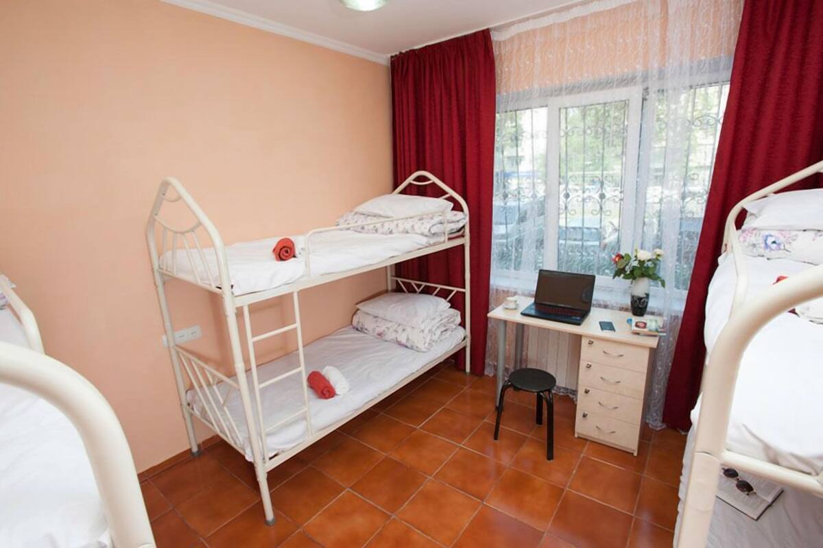 A dormitory room with bunk beds, a desk with a laptop, and a window with curtains.