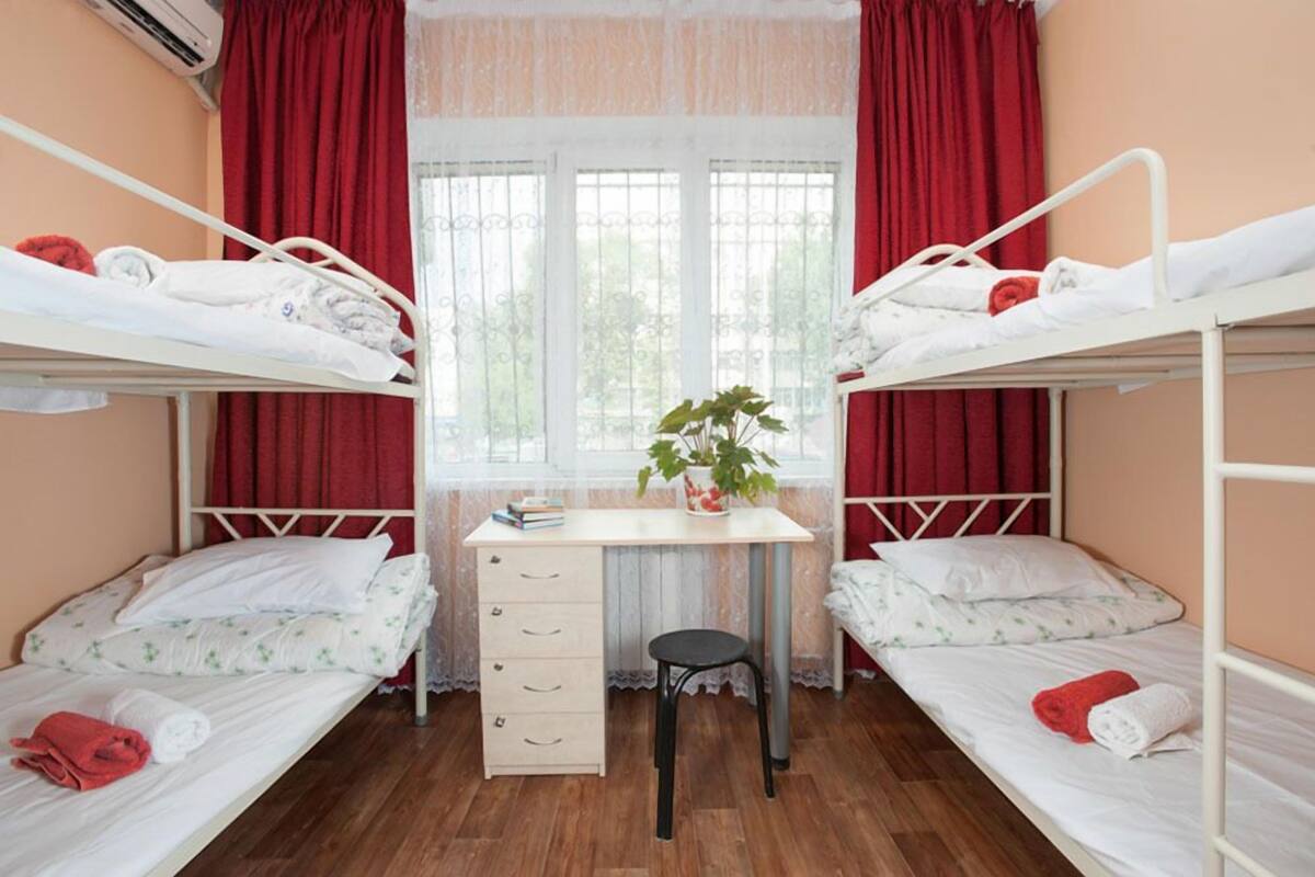 A dormitory room with two bunk beds, a desk, a chair, and a window with curtains.