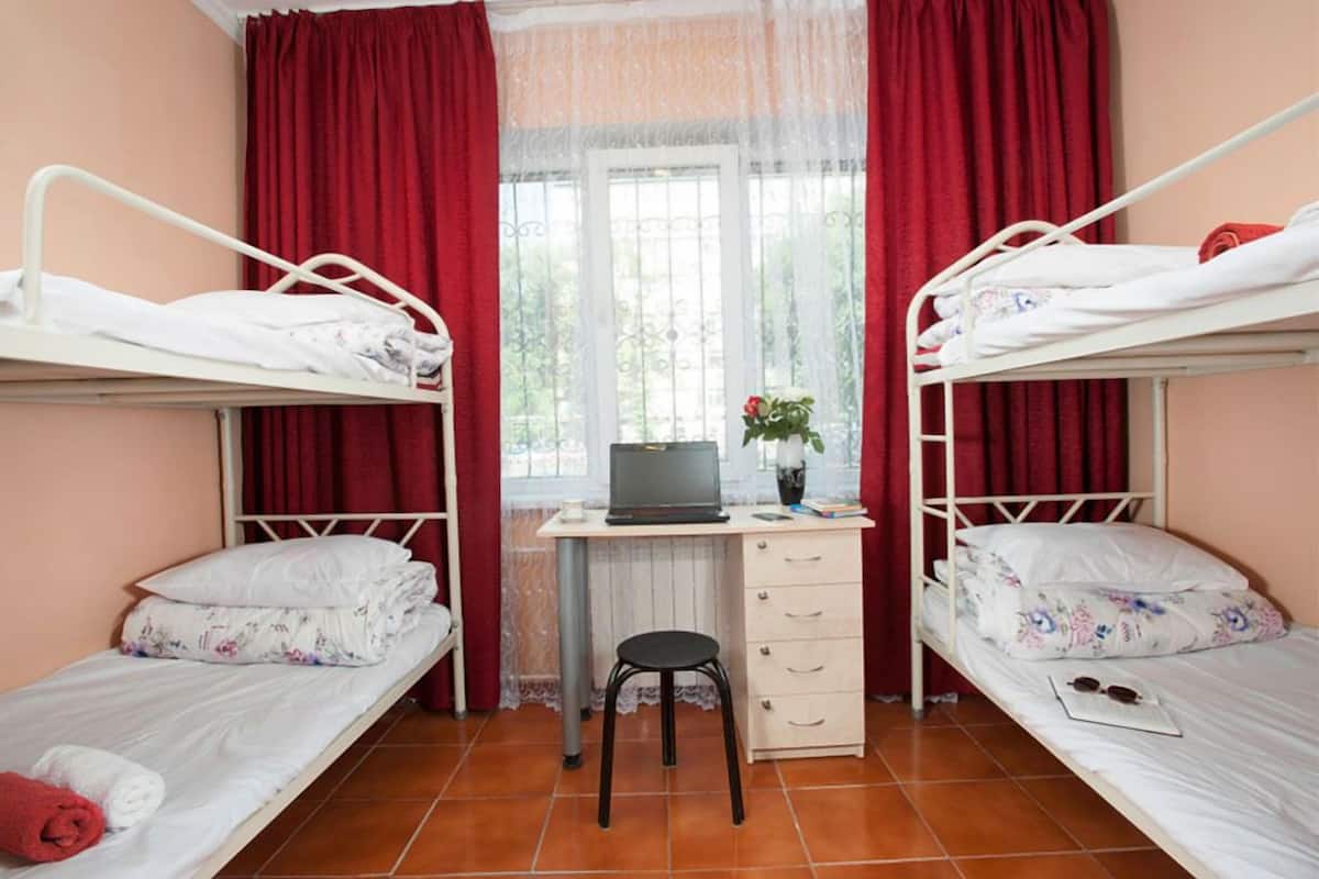 A dormitory room with two bunk beds, a desk with a laptop, a small stool, and a window with curtains.