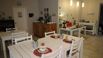 Un comedor con mesas y sillas blancas, una máquina de café y diversos electrodomésticos de cocina.