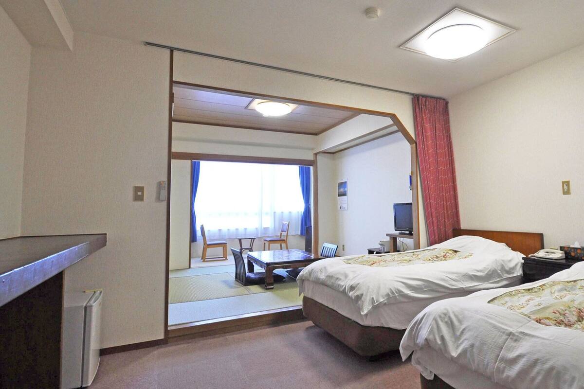 A hotel room with two beds, a desk, a chair, a television, and a view through an open door.