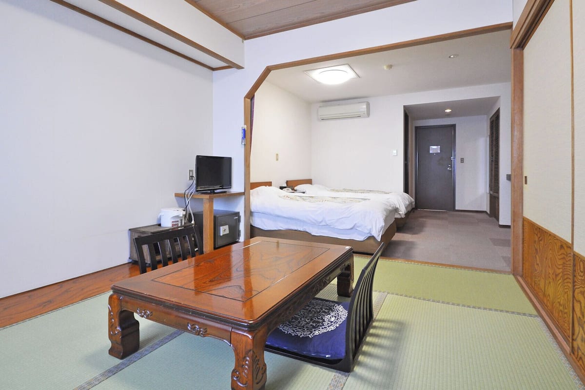 A hotel room with a wooden table, two beds, a television, and an air conditioning unit.