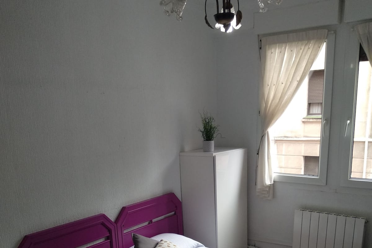 Un dormitorio con cama, mesita de noche, cómoda, ventana con cortinas y radiador.