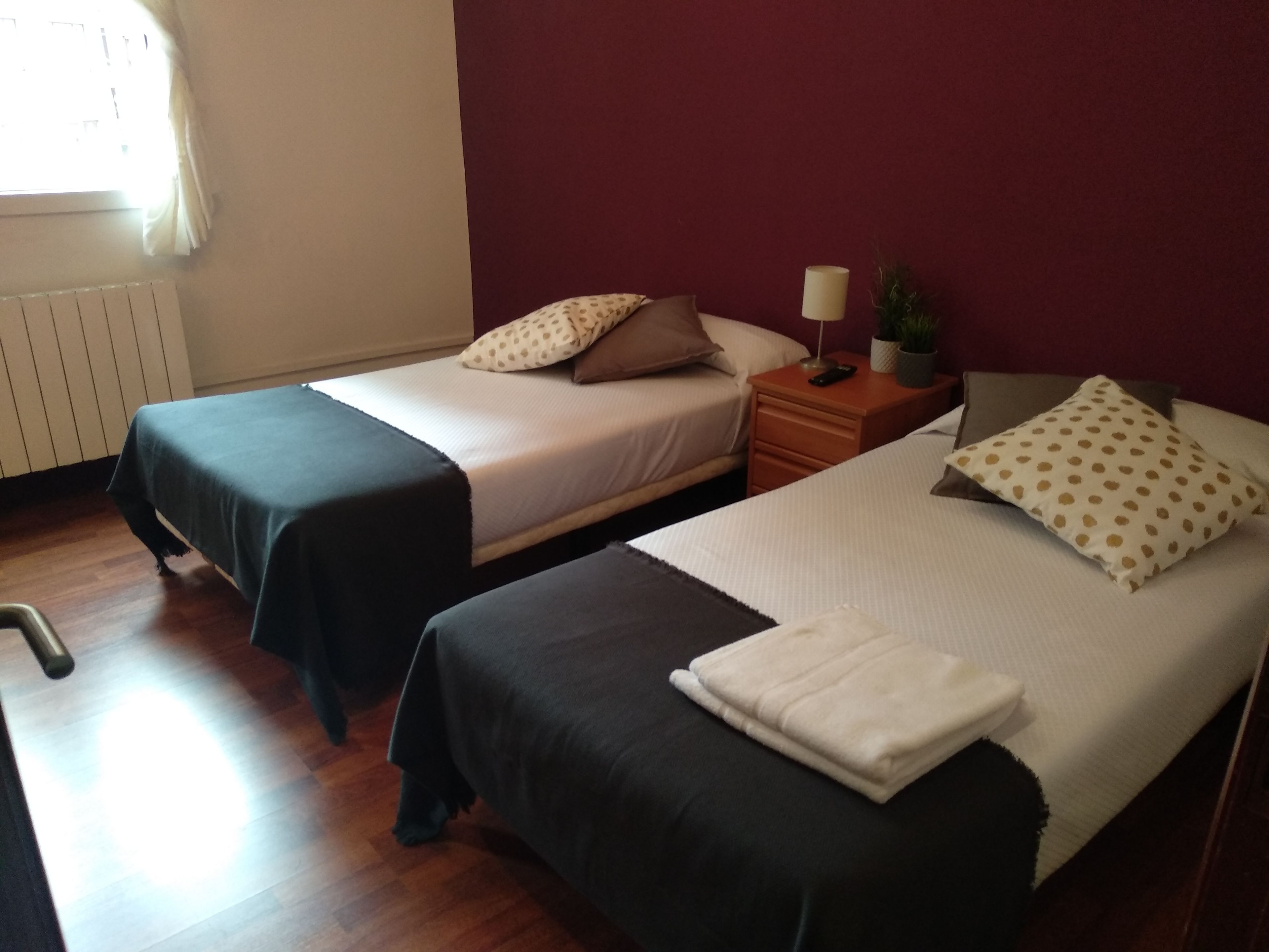 A room with two beds, each with a different bedspread, a nightstand with a lamp, and a radiator on the wall.