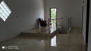 A spacious, well-lit room with a tiled floor, a chair, a small table, and a window providing a view of greenery outside. - Villa Pribadi Beunying 35 pax (Cipanas)